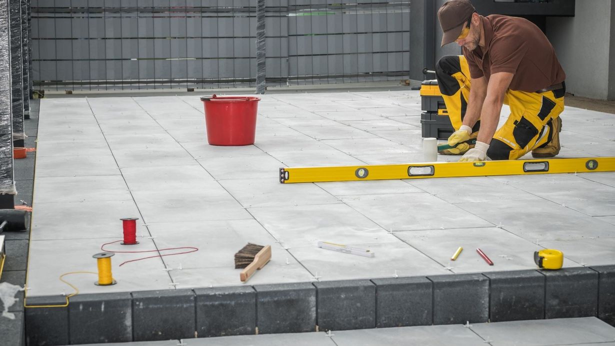 A construction worker lays paving stones on a patio, using a level.