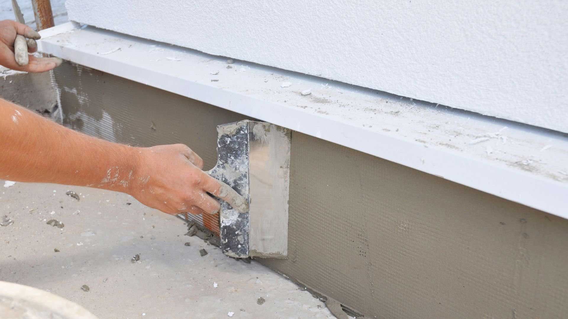 Person using a trowel to apply stucco to a building's foundation.