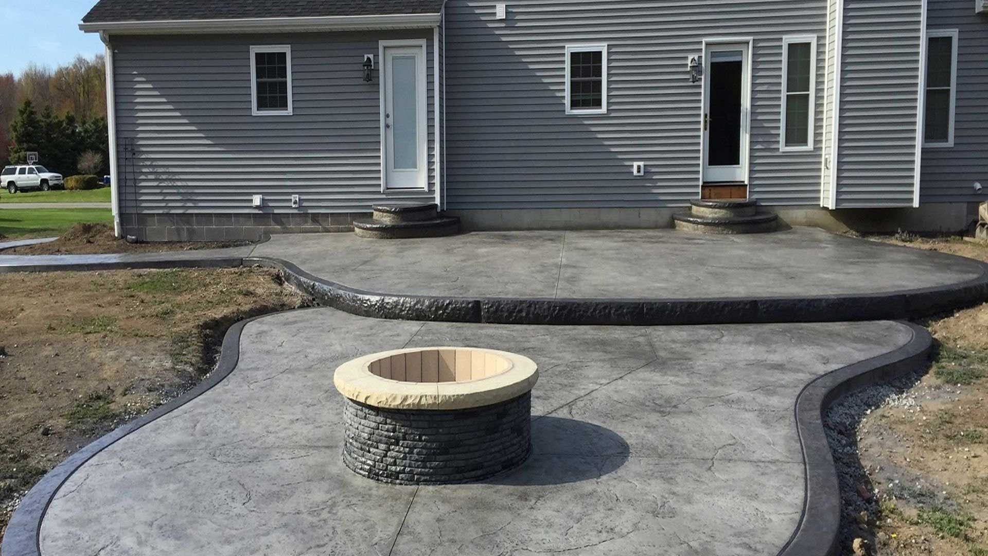 Concrete patio with fire pit, bordered by dark edging, in front of a gray-sided house with white doors.