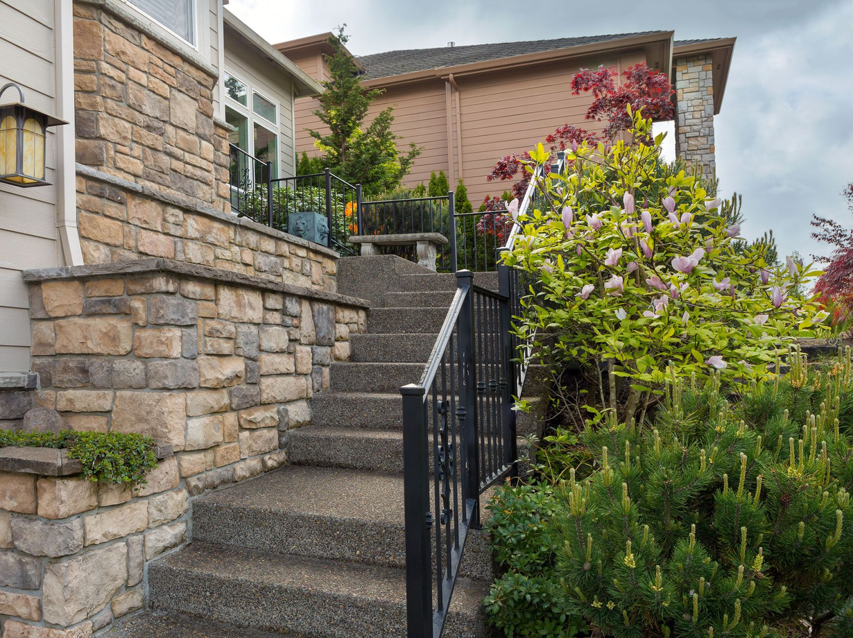 Stone steps lead up to a house entrance with stonework and a black railing, greenery on either side.