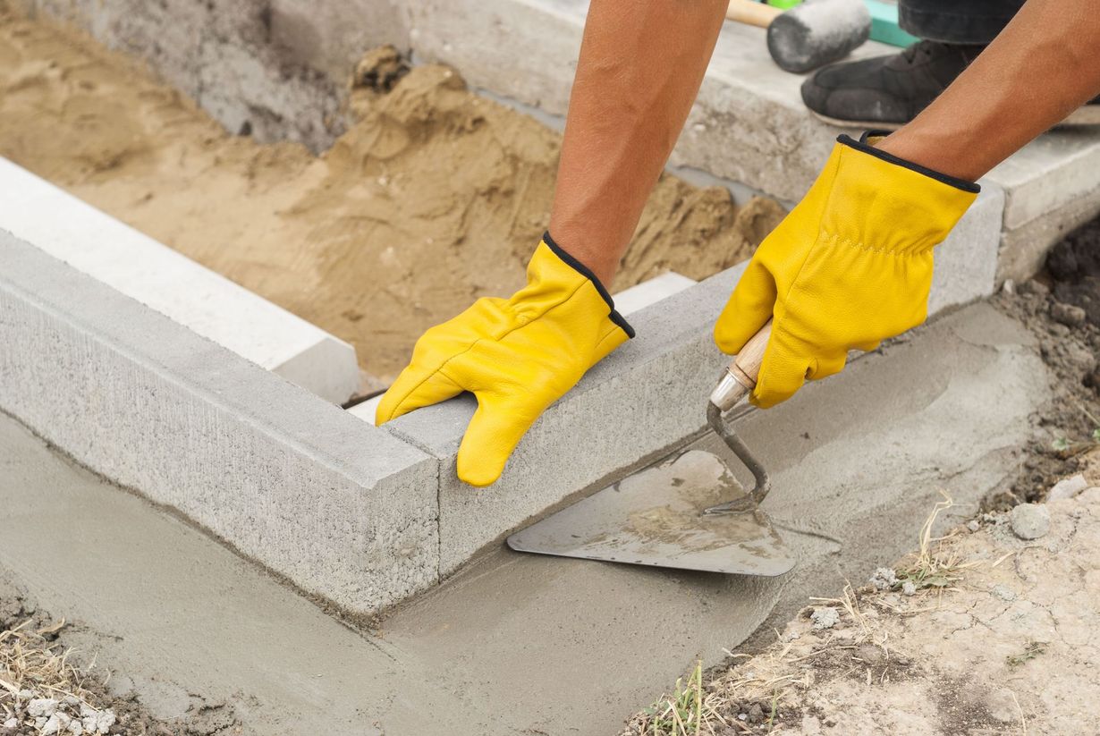 Person in yellow gloves using a trowel to lay a concrete block into mortar. Outdoors, setting blocks.