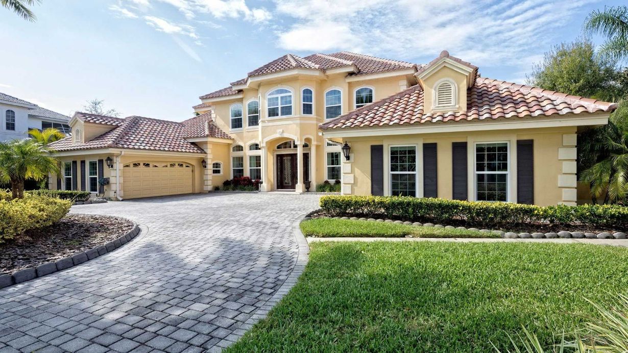 Yellow stucco mansion with a red-tiled roof, driveway, and lush green lawn.