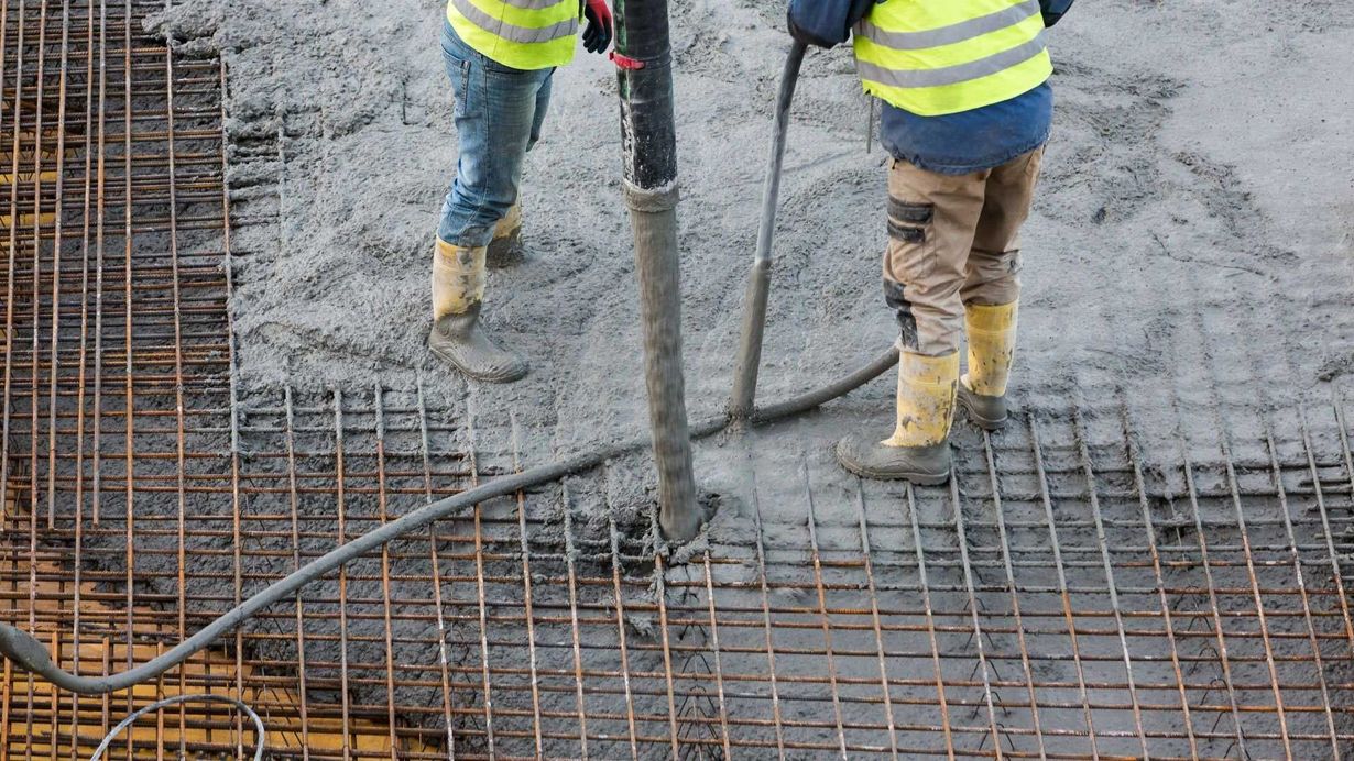 Construction workers pouring concrete over rebar grid wearing yellow vests and boots.