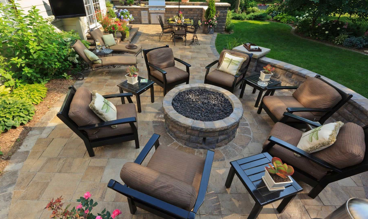Outdoor patio with a fire pit surrounded by seating, paved with stone, and a lush green lawn.