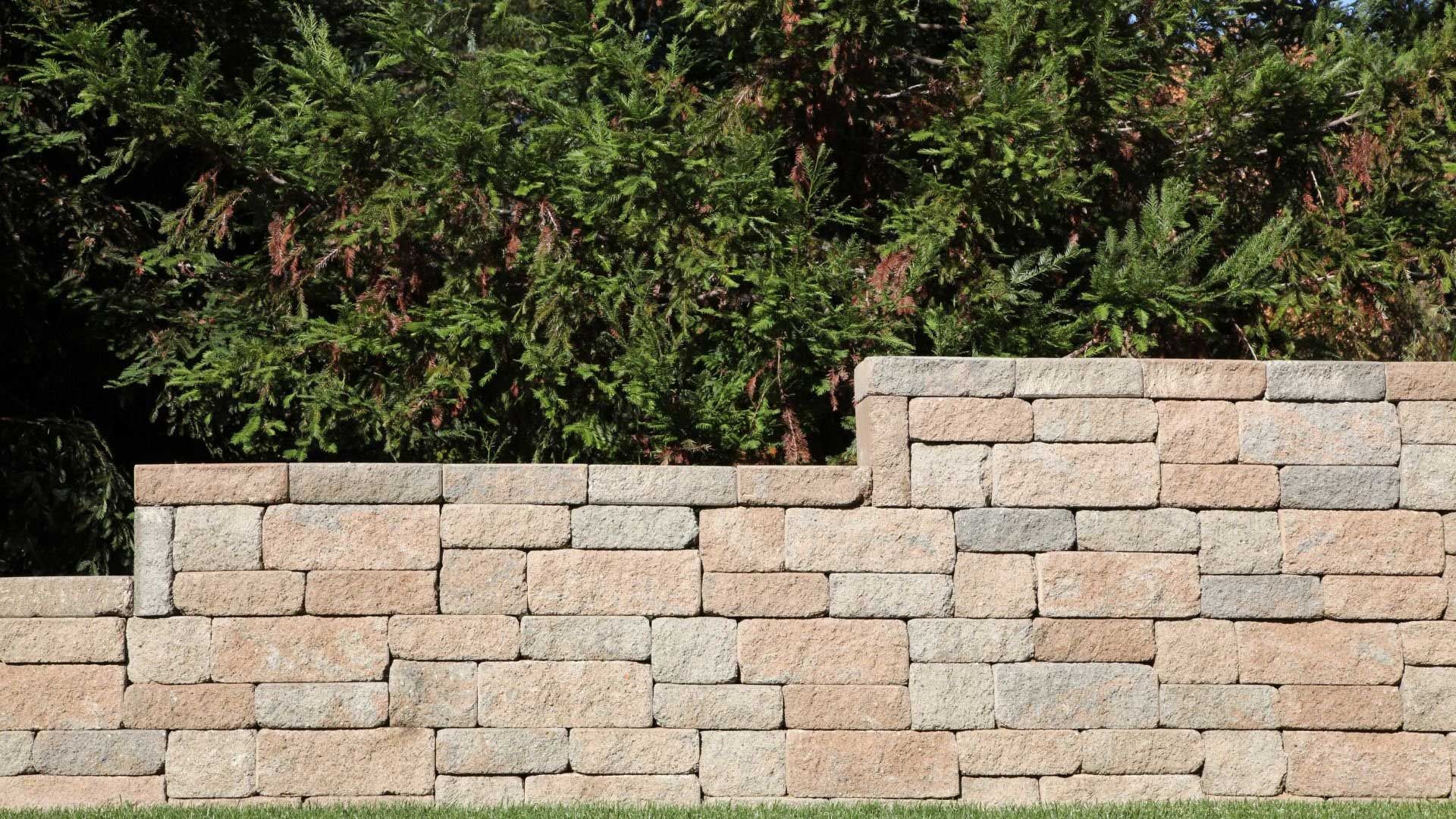 Stone retaining wall in front of green hedge. Wall is tan and gray with a step down.