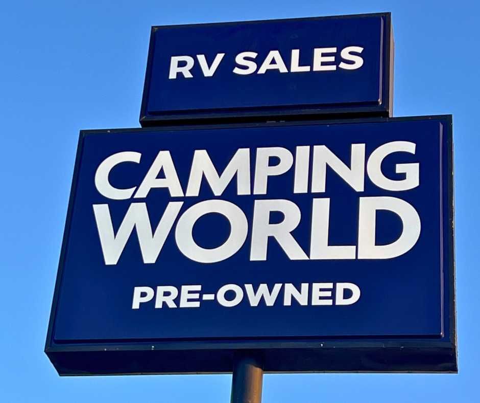 A blue rectangular sign against a clear sky reads RV SALES above CAMPING WORLD and PRE-OWNED in white text
