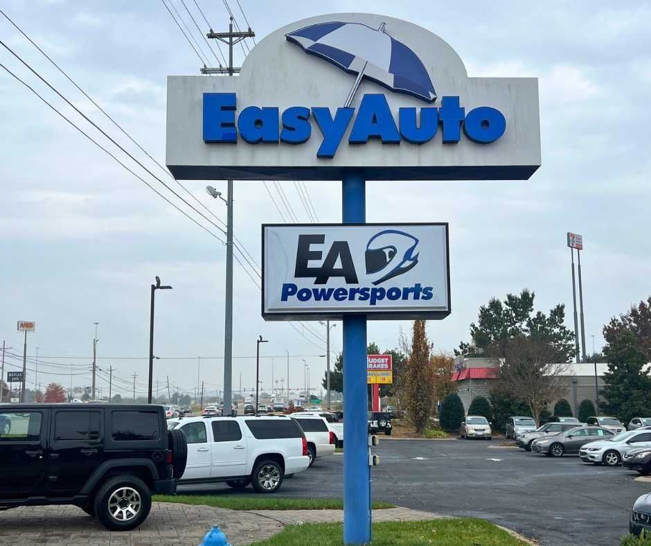 A sign for EasyAuto and EA Powersports, featuring a blue and white umbrella logo, standing in a car dealership parking lot
