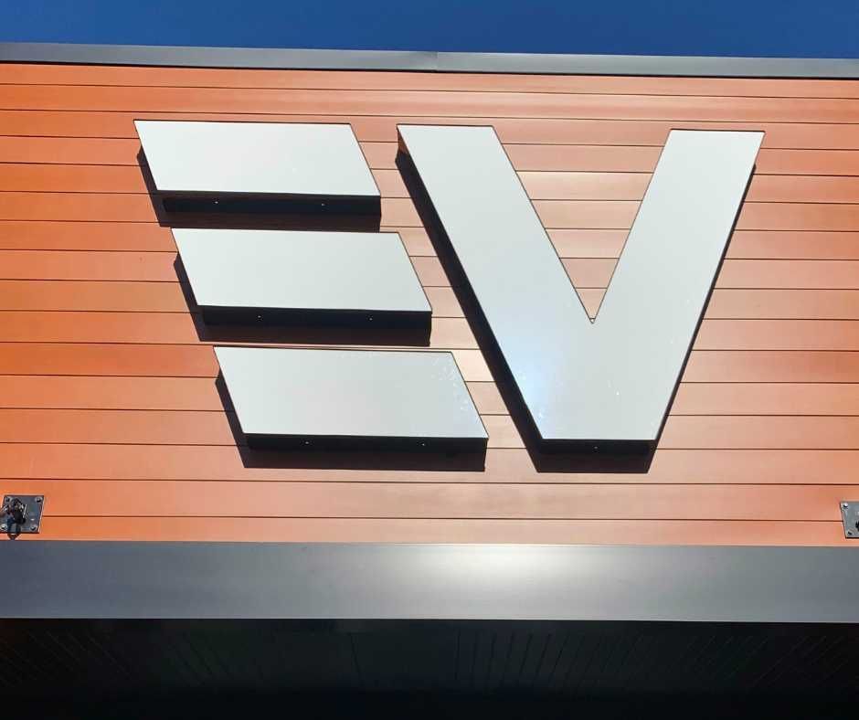 A silver-colored EV logo mounted on a wall with horizontal, light-brown, wood-like panels against a blue sky