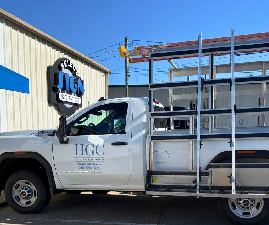 A white HGC service truck with a glass rack and ladder is parked in front of a building with a SELECT SIGN SERVICE sign