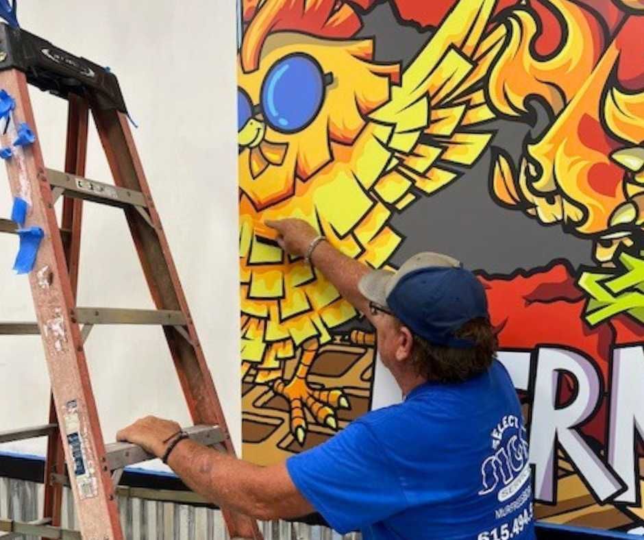 A person on a ladder paints a mural featuring a yellow cartoon bird wearing blue glasses next to large flames