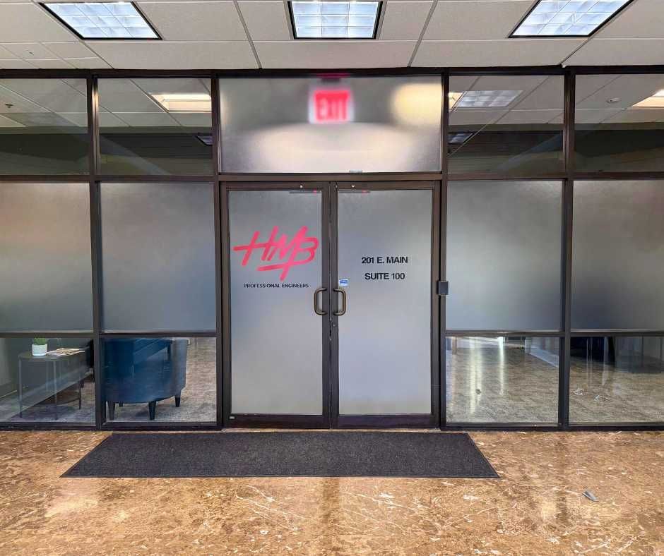 Glass office entrance with HMB logo on the frosted doors, located in a tiled hallway with a black door mat
