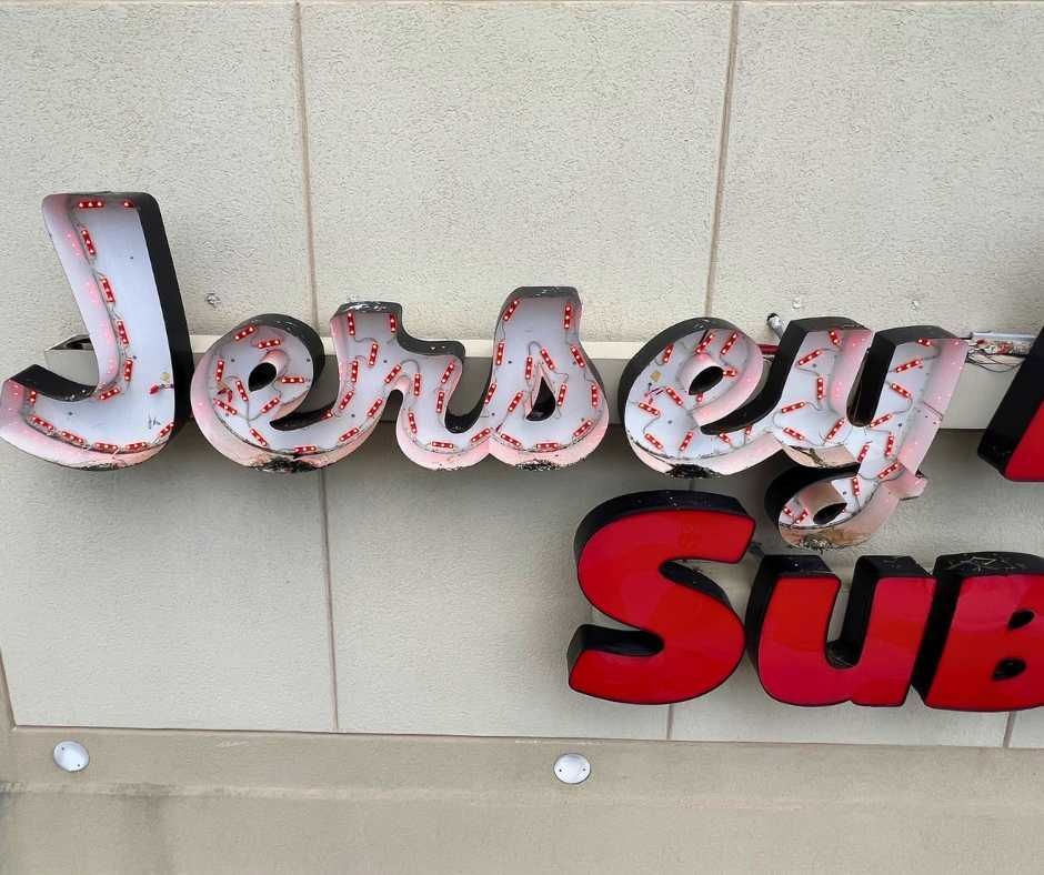 A Jersey Mike's Subs exterior sign on a beige wall, featuring white light-up lettering and a red SUBS sign below it