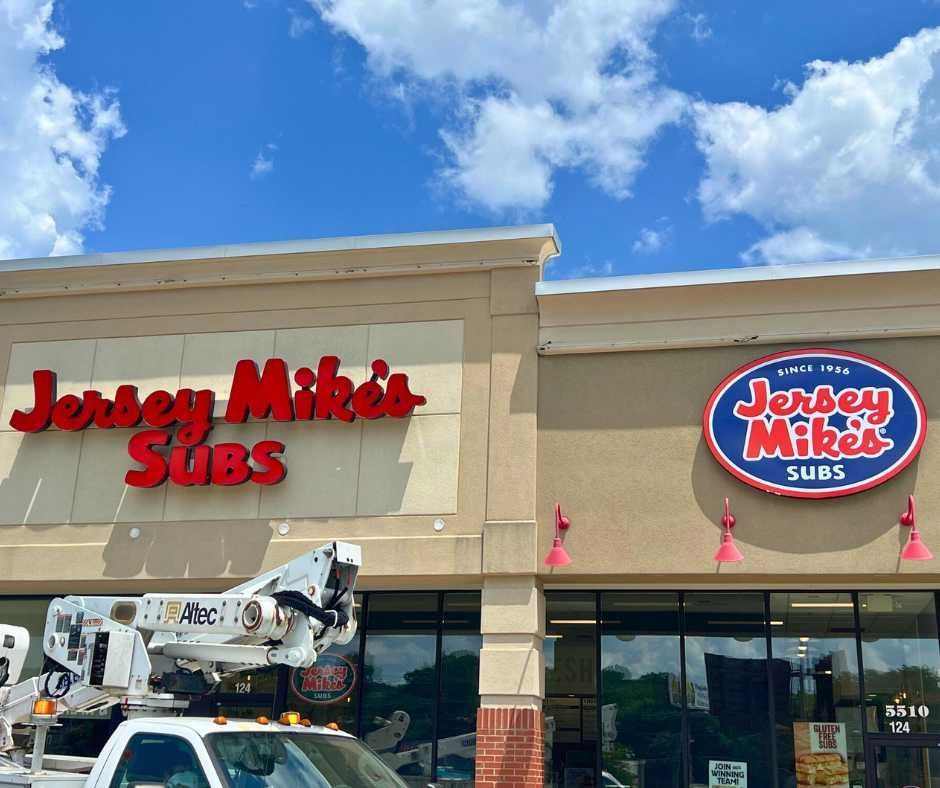 A Jersey Mike’s Subs storefront with a cherry picker truck parked in front, beneath a blue sky with white clouds