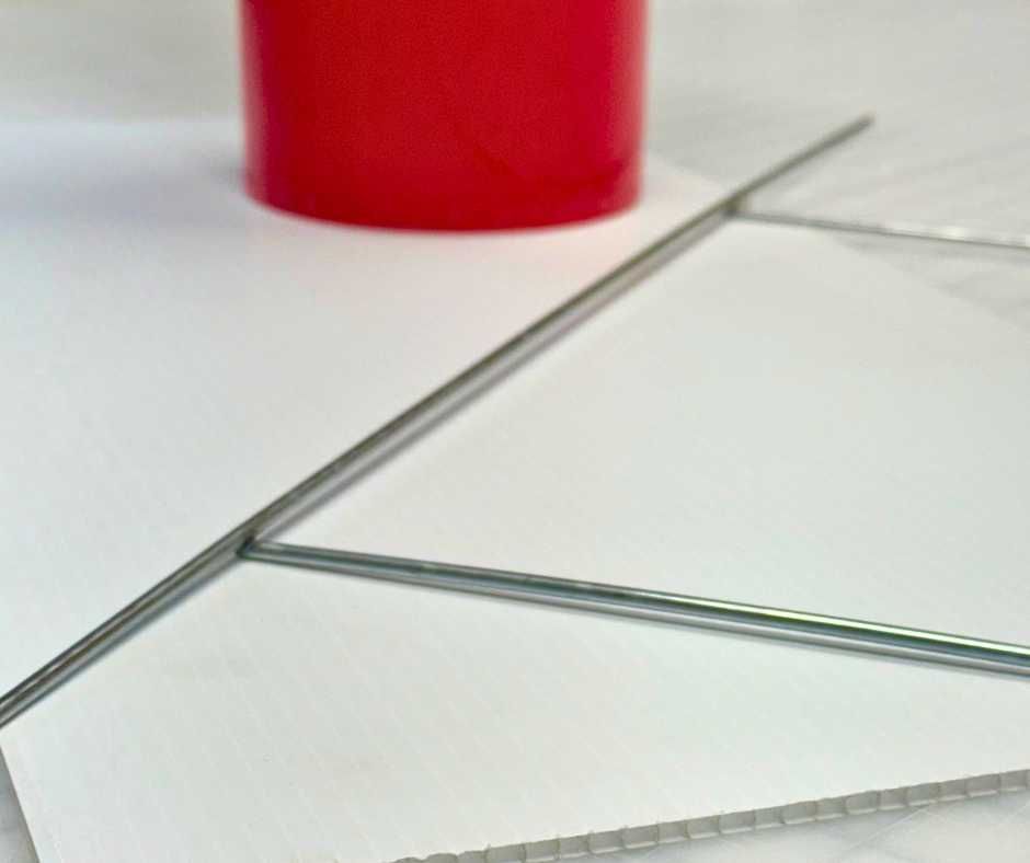 A red plastic tube resting on a white corrugated board beside thin metal rods arranged in a Y-shape