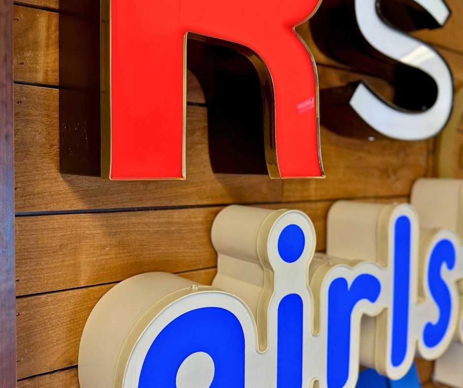 Close-up of bright, 3D channel letters—a red R above blue girls—mounted on a slatted, brown wooden wall
