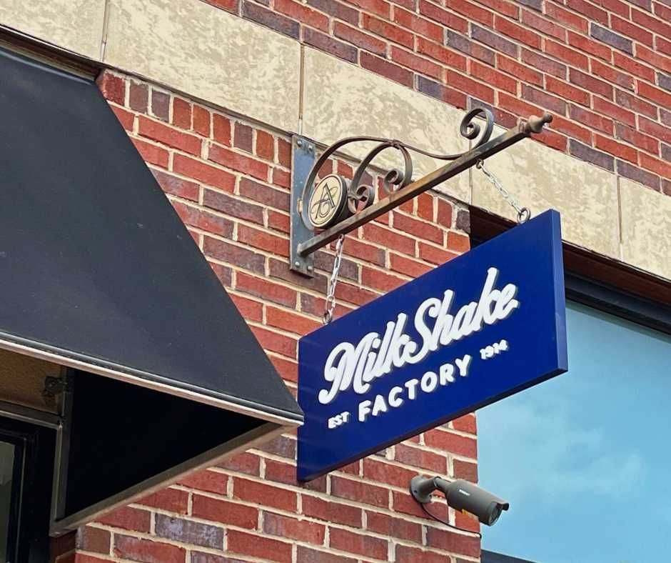 A blue, rectangular MilkShake Factory sign hangs from a decorative black metal bracket on a red brick wall