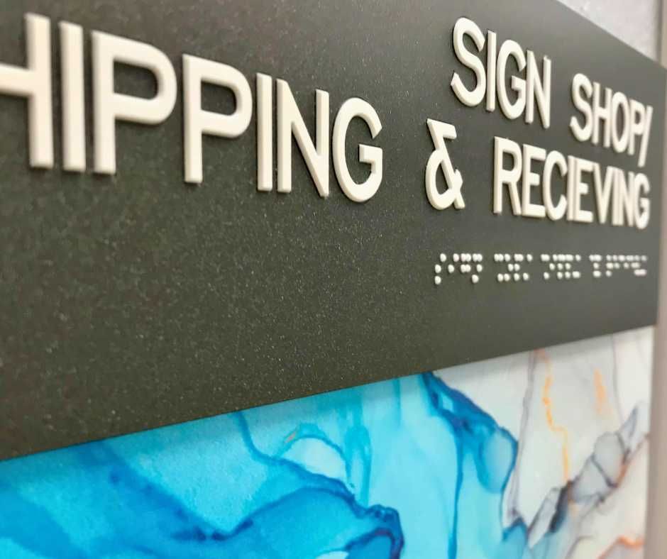 A black sign with white raised lettering reading SHIPPING, SIGN SHOP, and RECEIVING above Braille