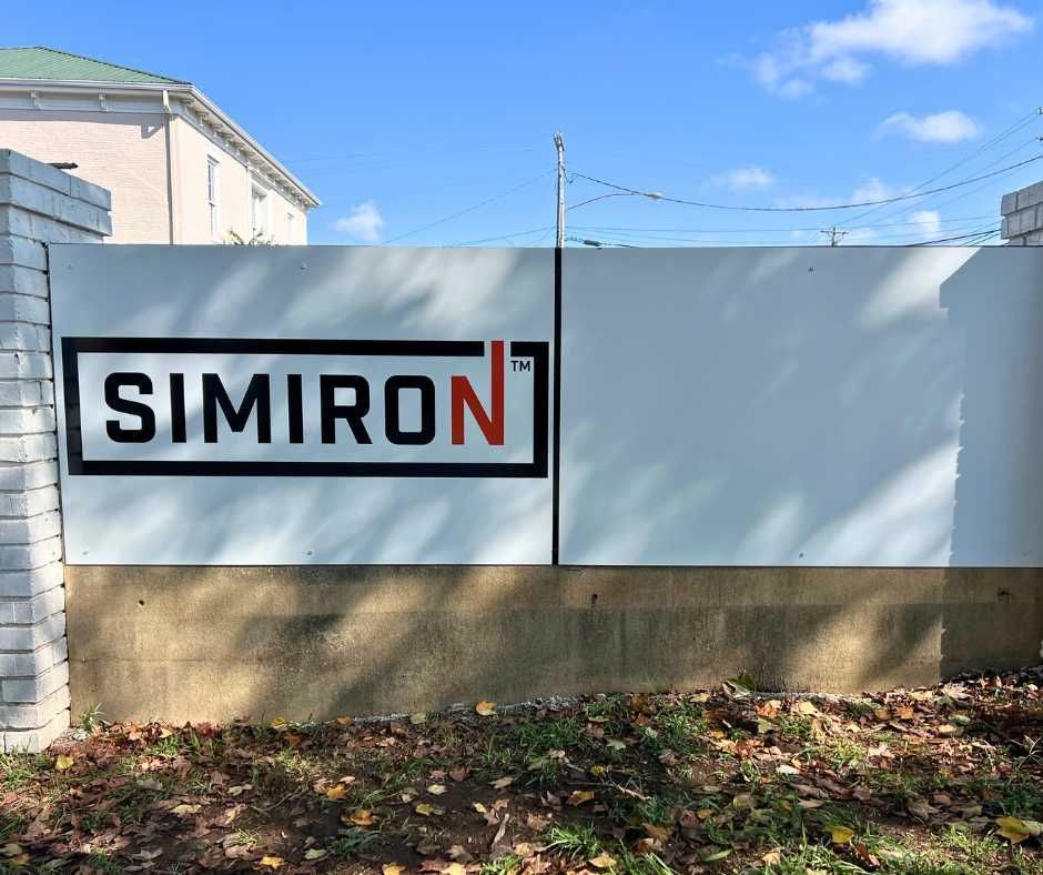 A white wall sign with the black and orange text SIMIRON inside a black rectangular frame, set in an outdoor area