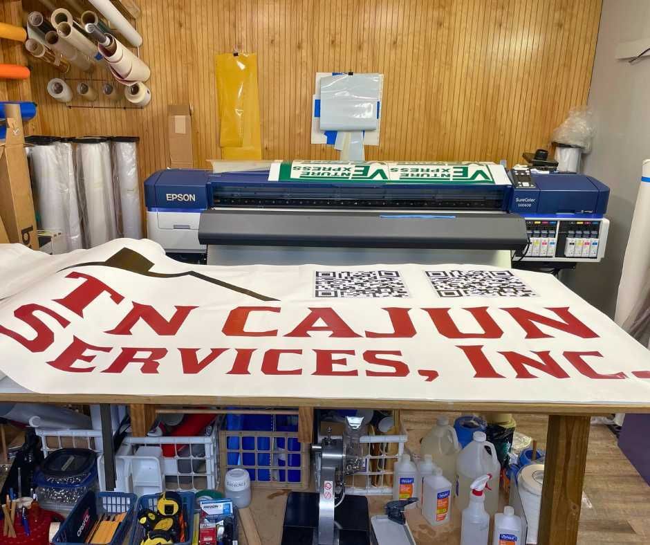 A sign for TN Cajun Services, Inc. with two QR codes resting on a table in front of a large format printer