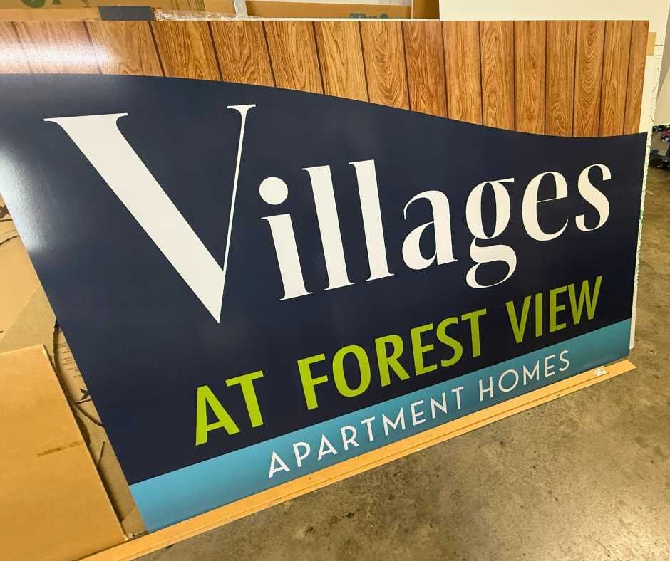 Sign reading Villages AT FOREST VIEW APARTMENT HOMES against a wood-paneled background in a workshop setting