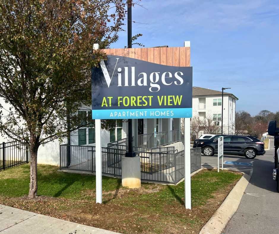 A sign for Villages at Forest View Apartment Homes, featuring dark blue and light blue text on a white pole in a park