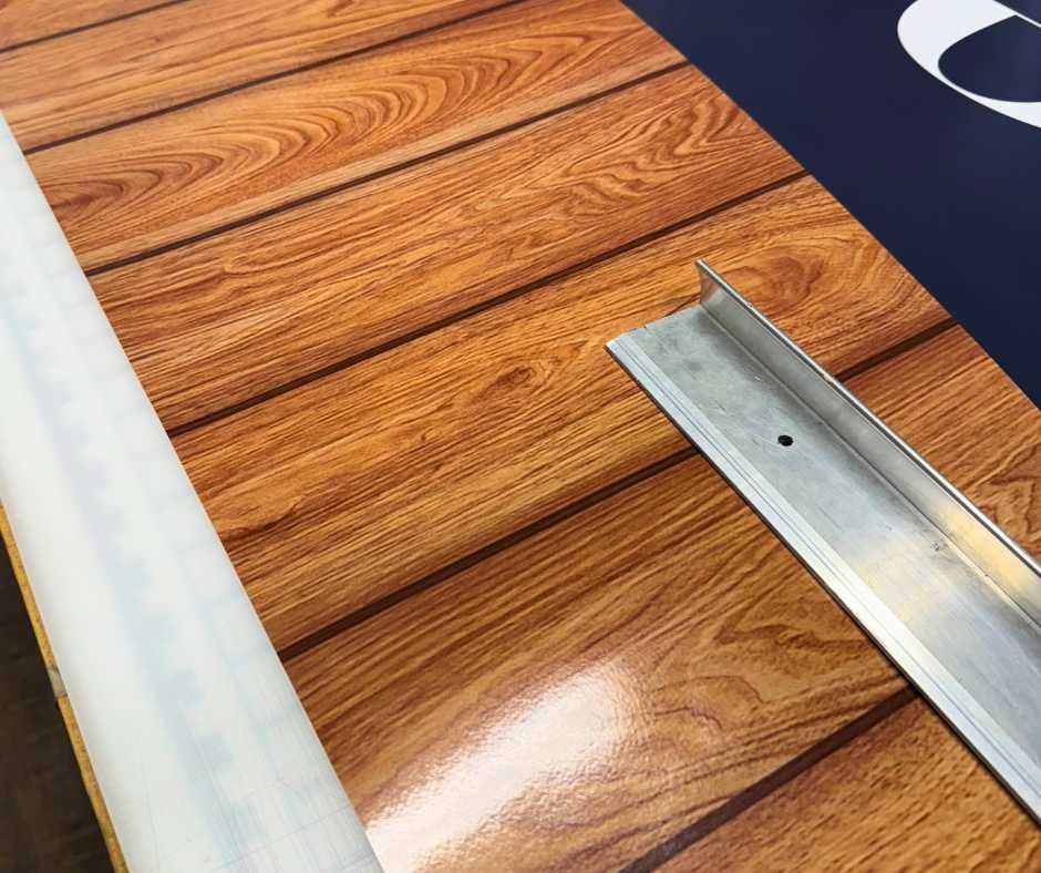 A metallic L-bracket sits on a polished, wood-grain patterned surface next to a white edge, with blue signage above