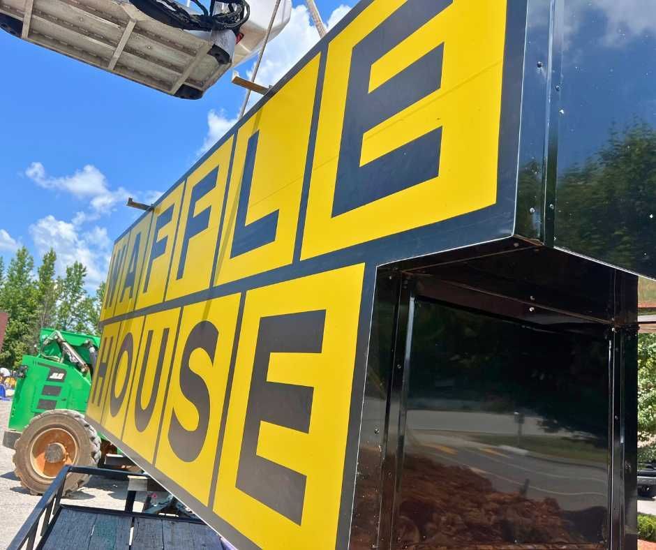 A large, yellow Waffle House sign is being lifted by a crane near a green construction vehicle under a blue sky