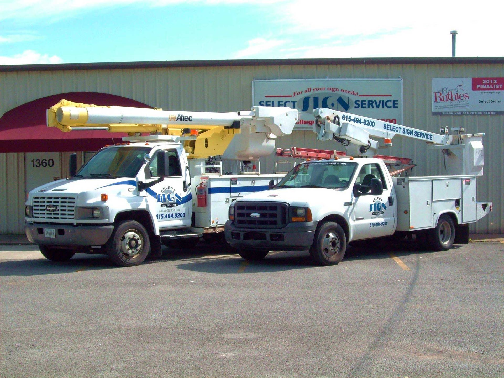Two white service trucks parked outside a building with a red awning; one truck has a raised boom lift.