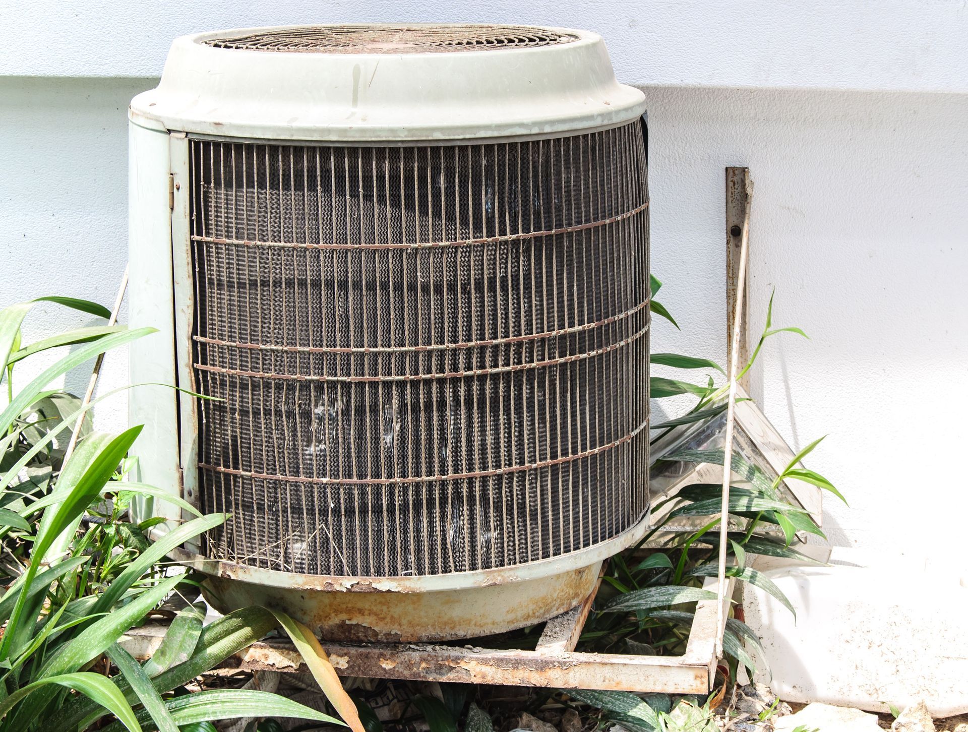 Outdoor air conditioner unit with a dirty, rusted grille beside a wall and plants
