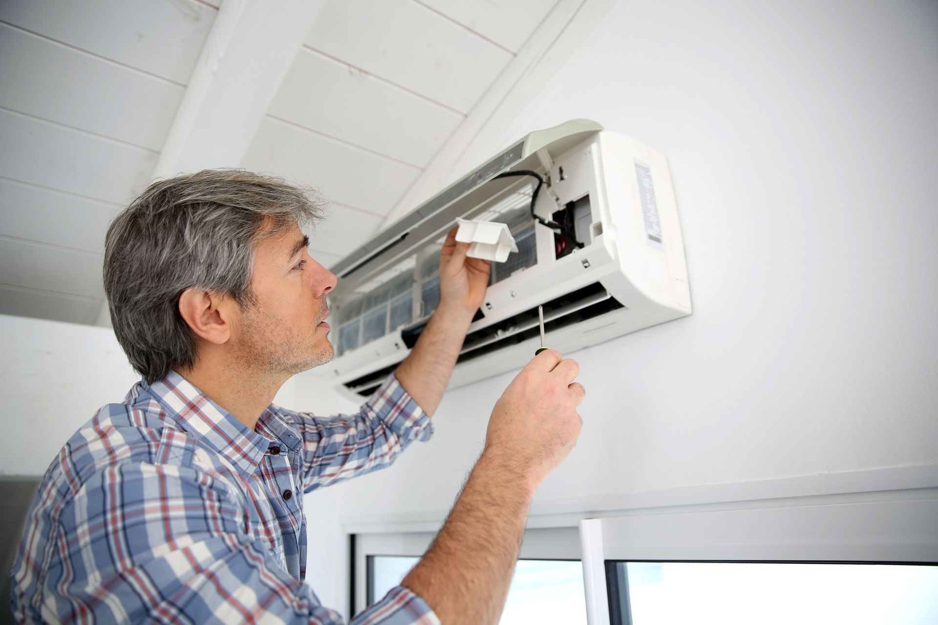 Man cleaning an air conditioner unit on a white wall with a cloth and screwdriver