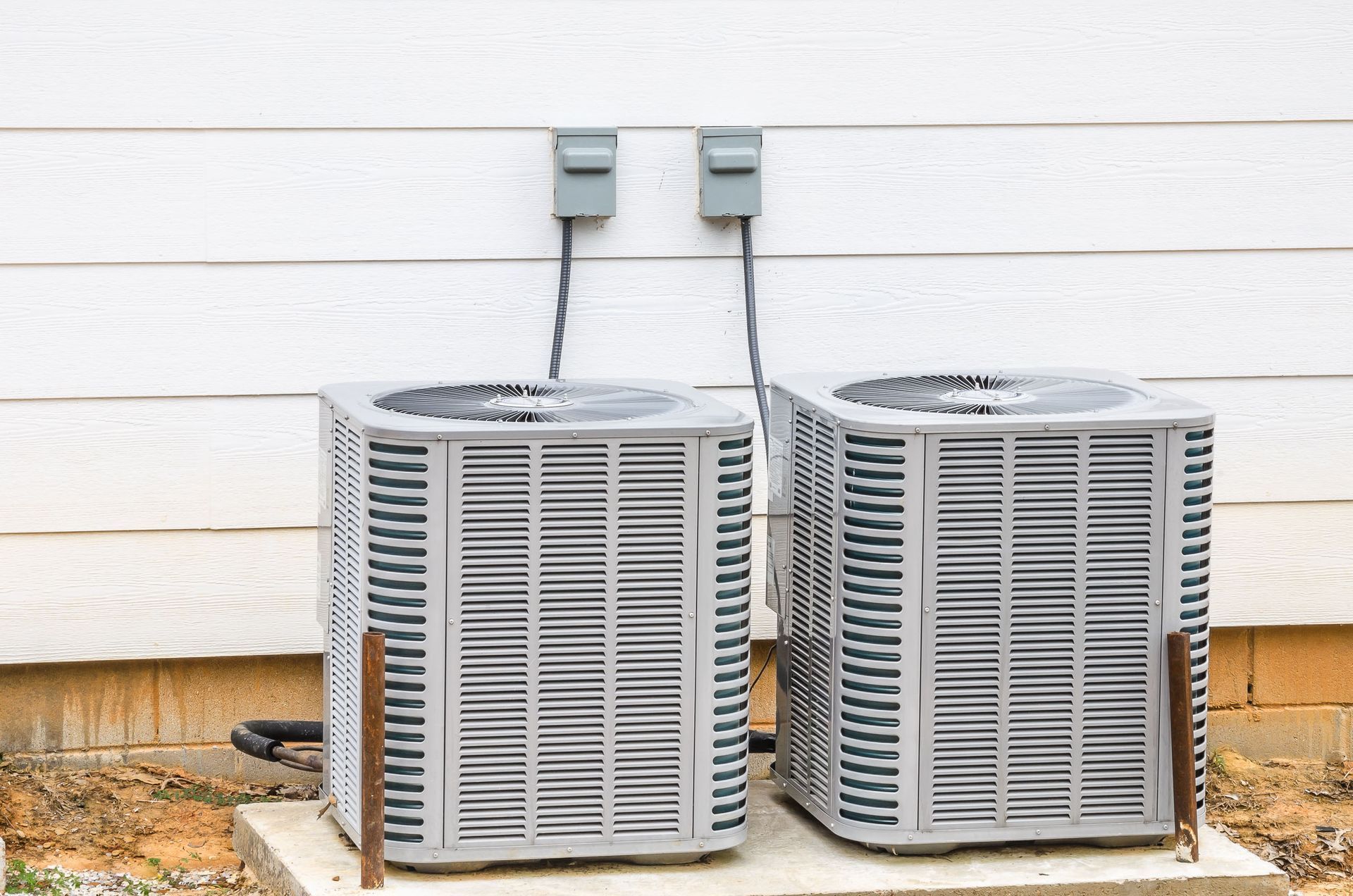 Two gray outdoor air conditioning units beside a white house wall with electrical boxes above