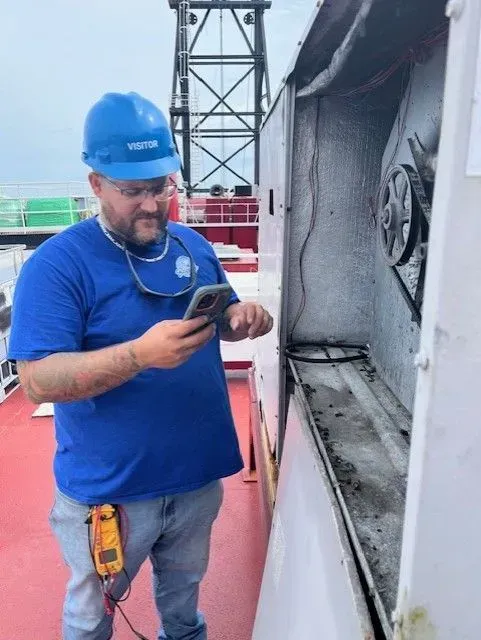Worker in blue hard hat and shirt checking a device beside industrial equipment on a ship deck