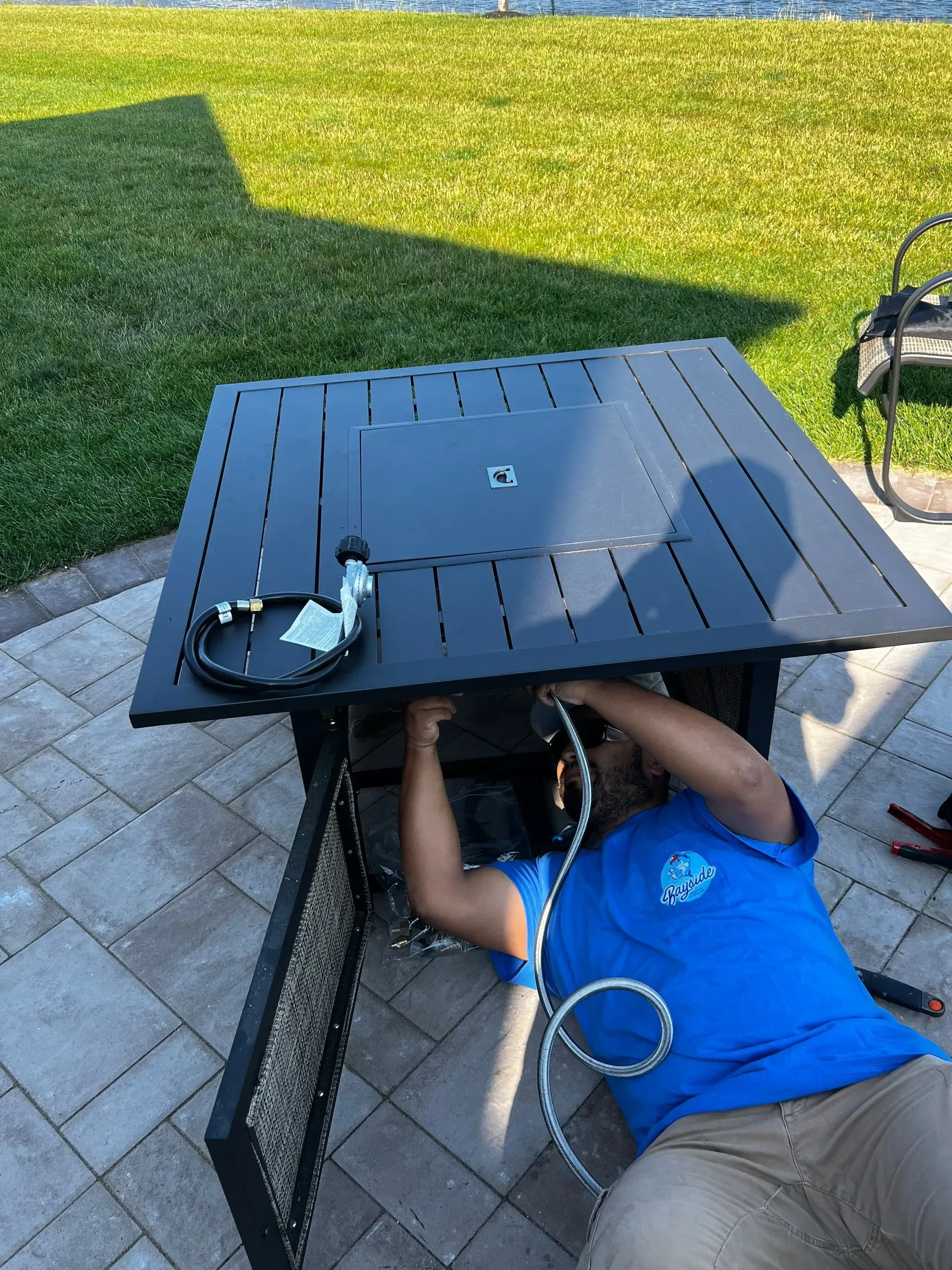 Person lying under a black patio table in a backyard, with a blue shirt and tools nearby