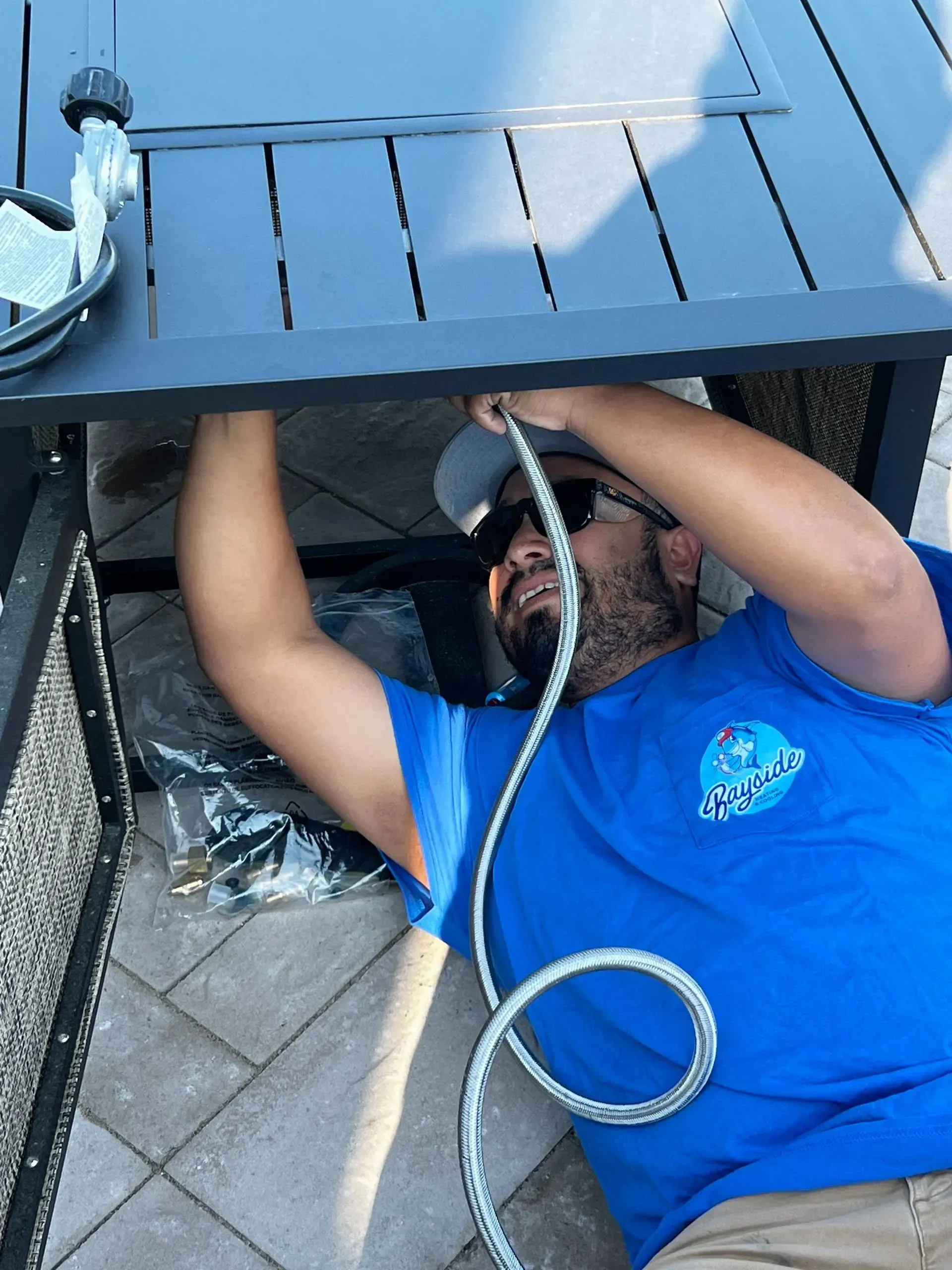 Technician in blue shirt working under a raised structure, reaching up with tools and a coiled cable.
