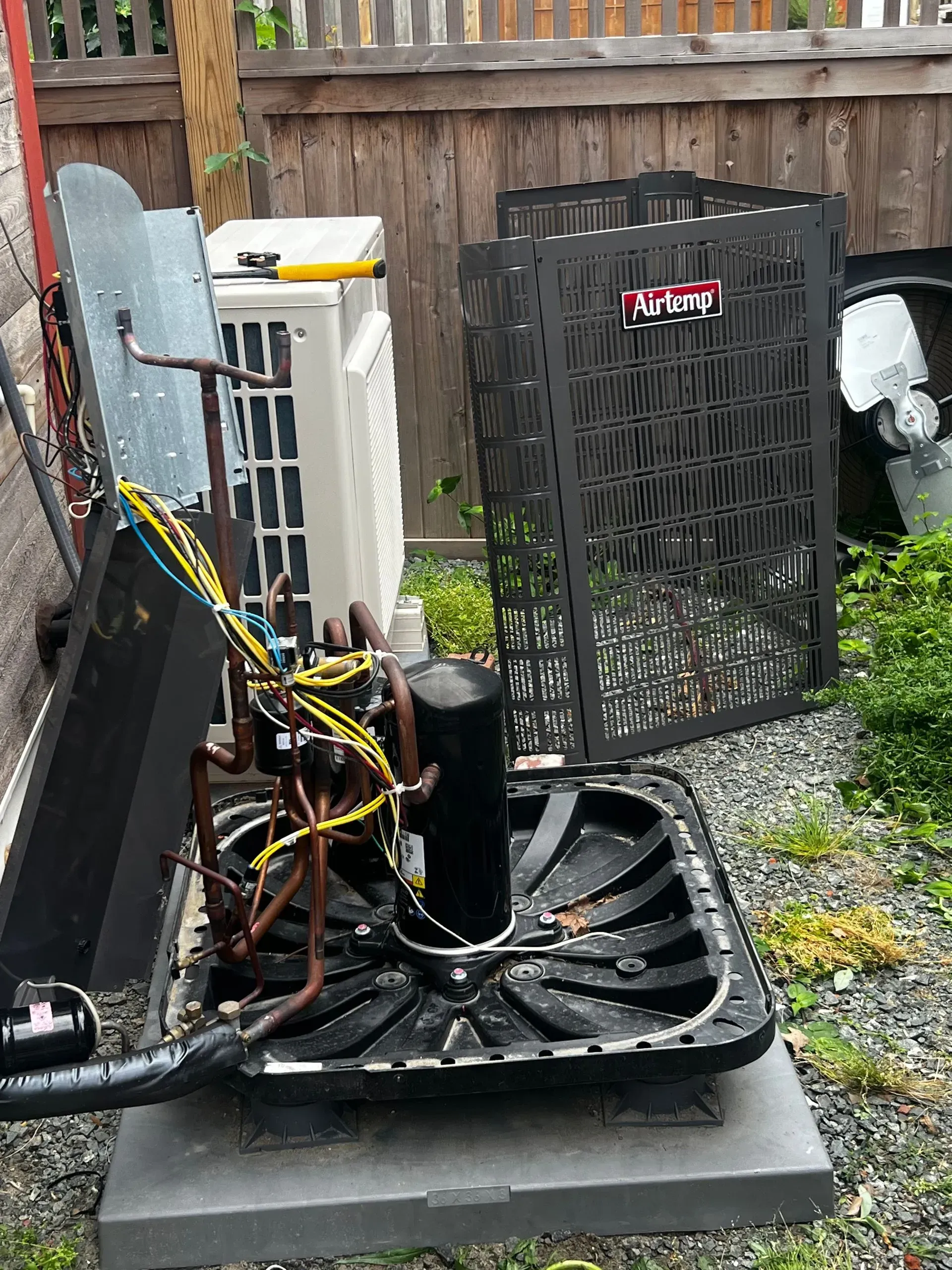 Outdoor utility equipment with exposed wires and a black fan unit beside an air conditioner in a fenced yard
