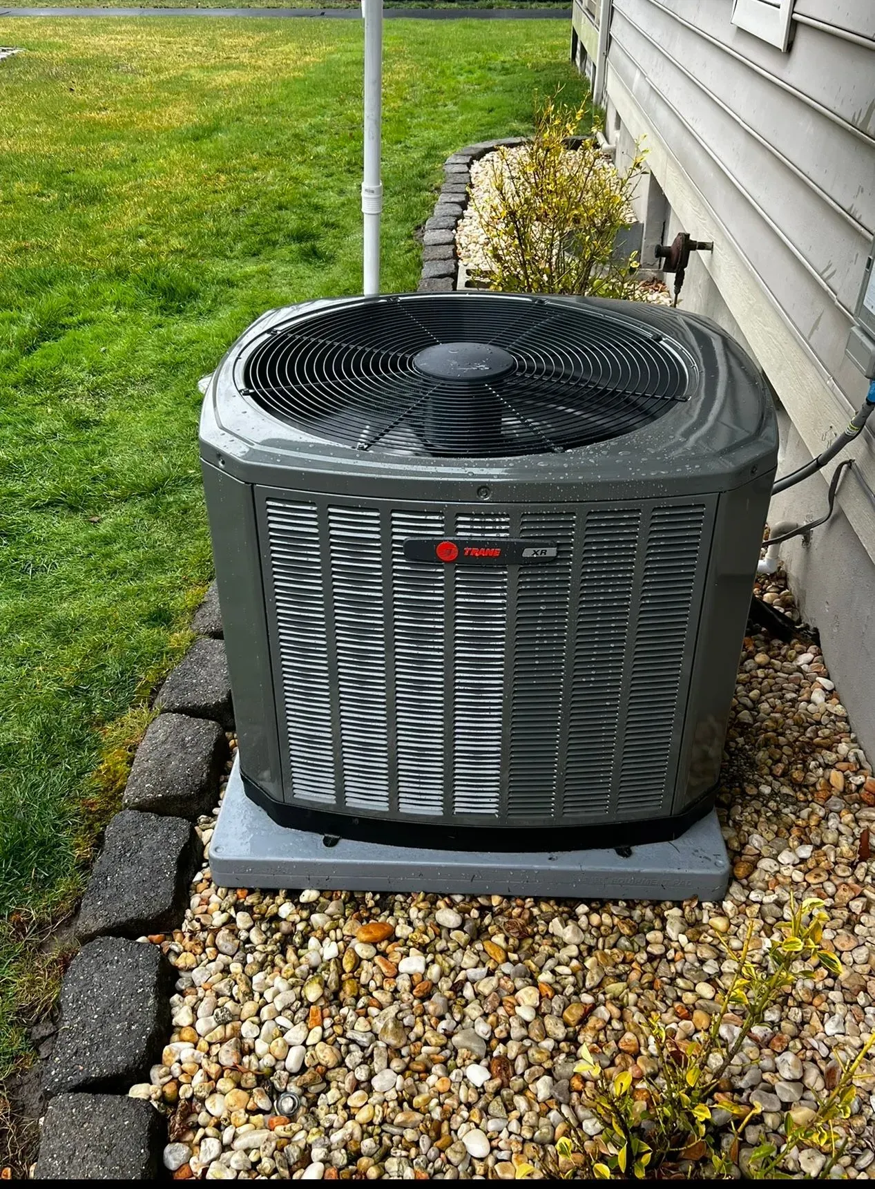 Outdoor air conditioner unit beside a house, with black top and gray grille on a gravel bed.
