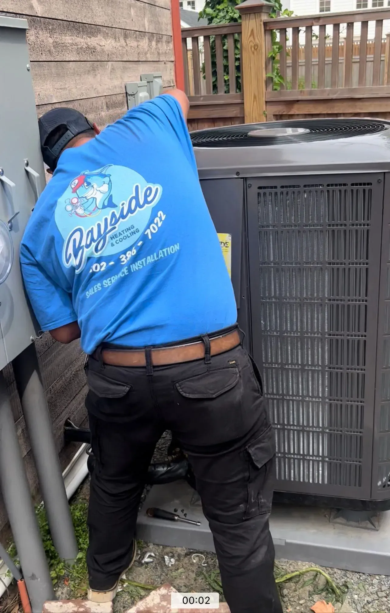 Technician in a blue shirt working on an outdoor HVAC unit beside a wooden deck.