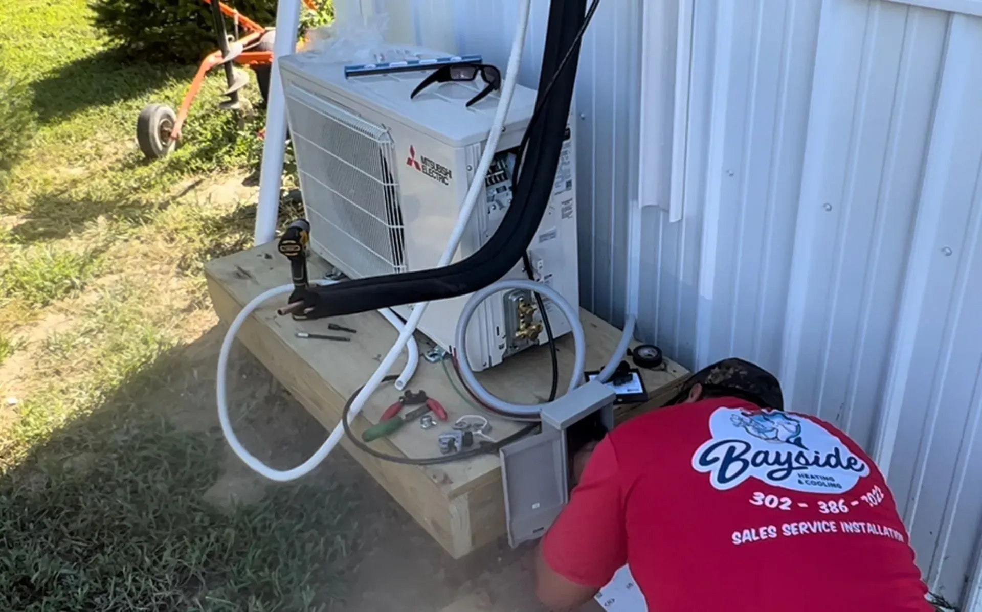 Outdoor utility equipment beside a white wall, with hoses and tools on the ground and a person in a red shirt kneeling.