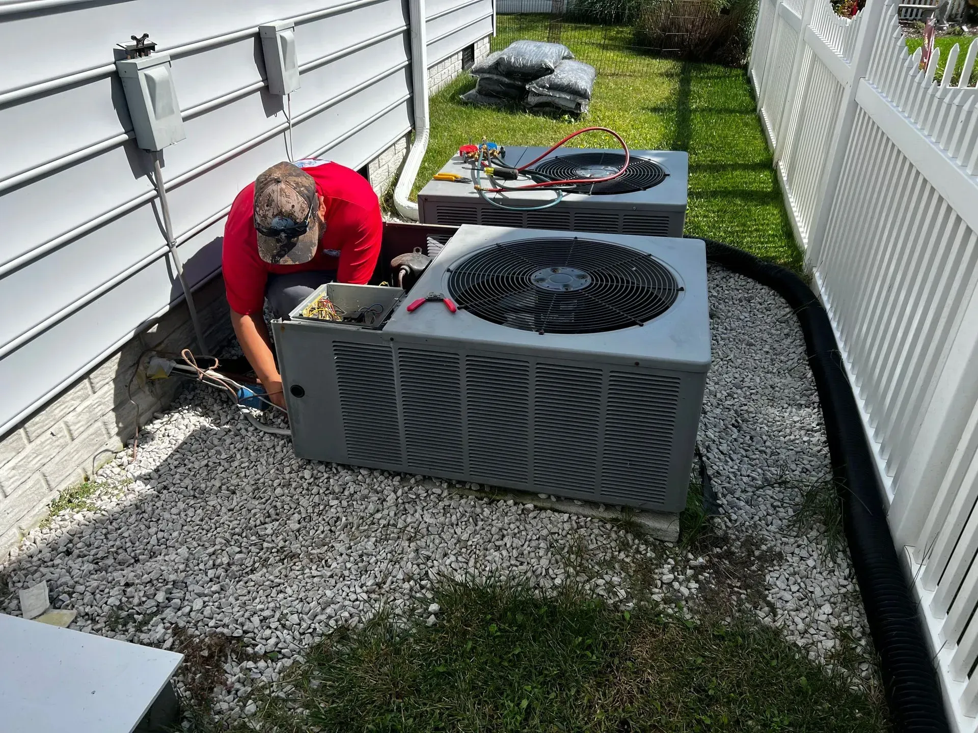 Technician kneeling beside a large outdoor air conditioner unit along a narrow gravel side yard.