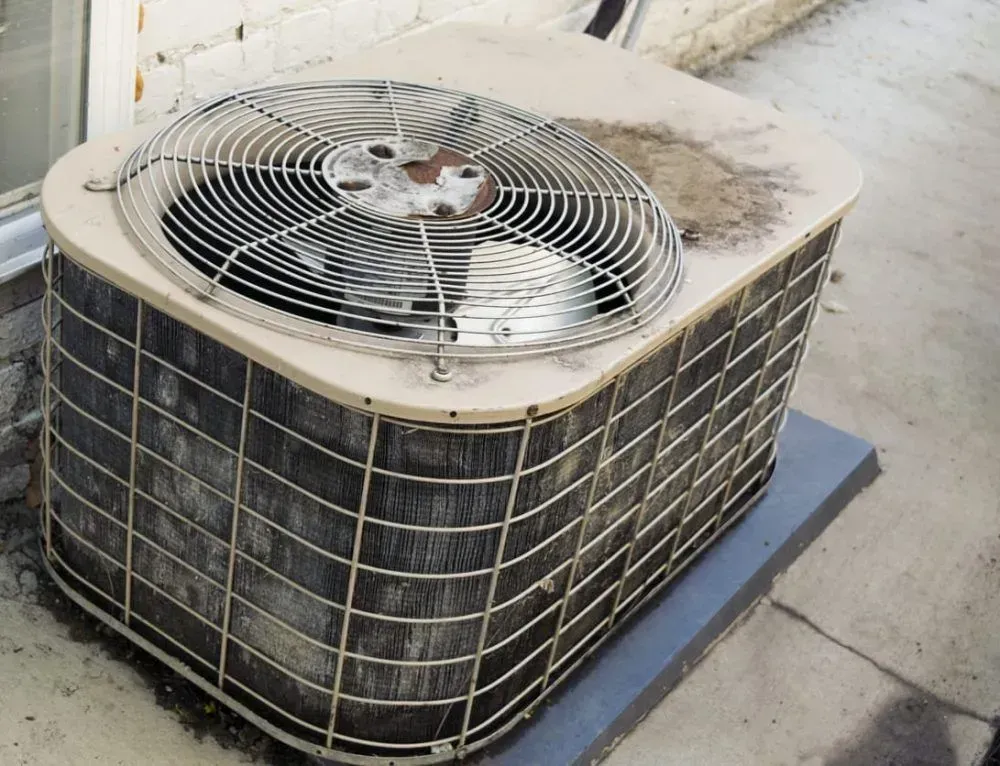 Dirty outdoor air conditioner unit on a concrete patio next to a wall.