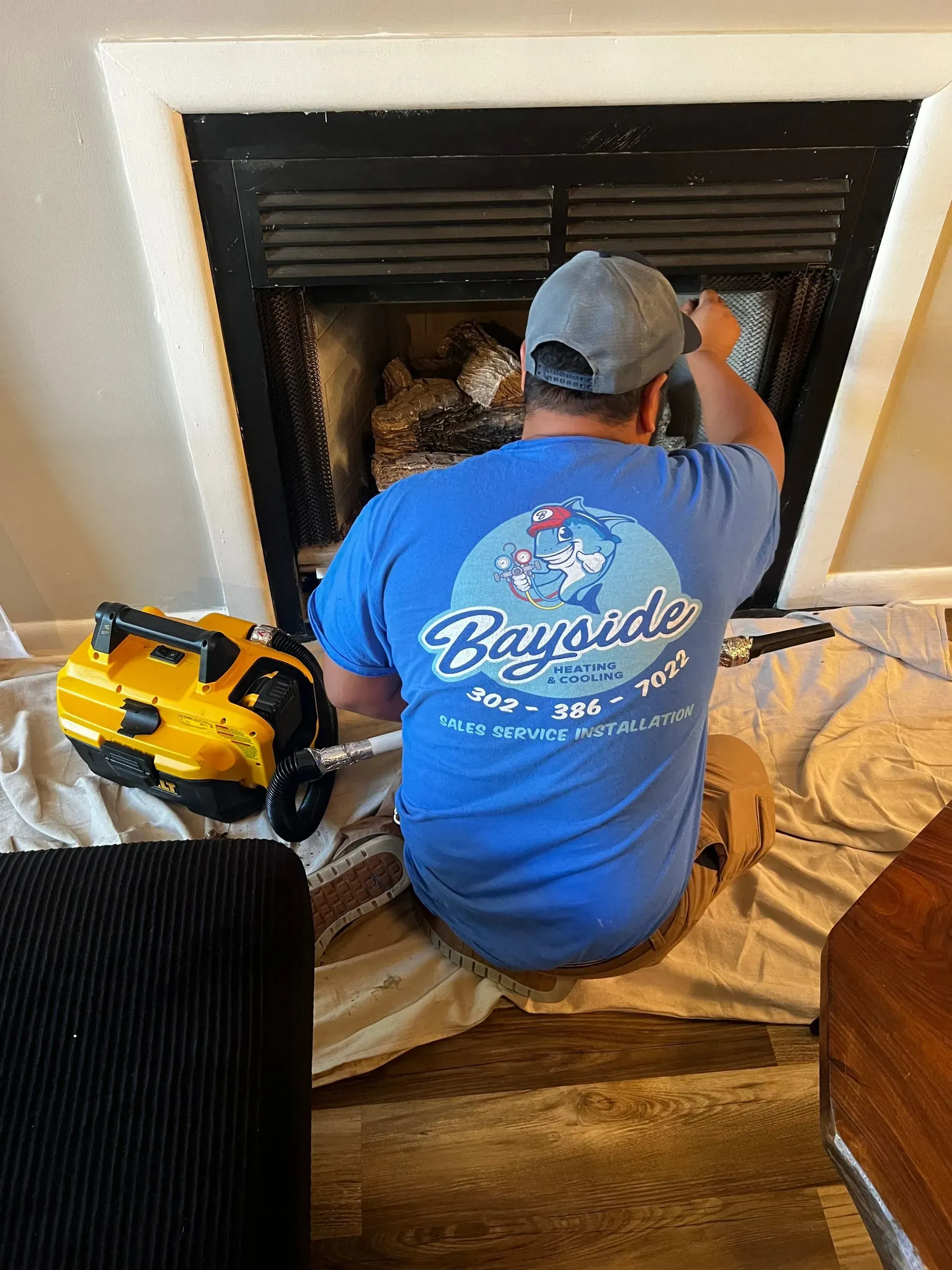Technician in blue shirt cleaning a fireplace, with a yellow tool bag on the floor nearby.