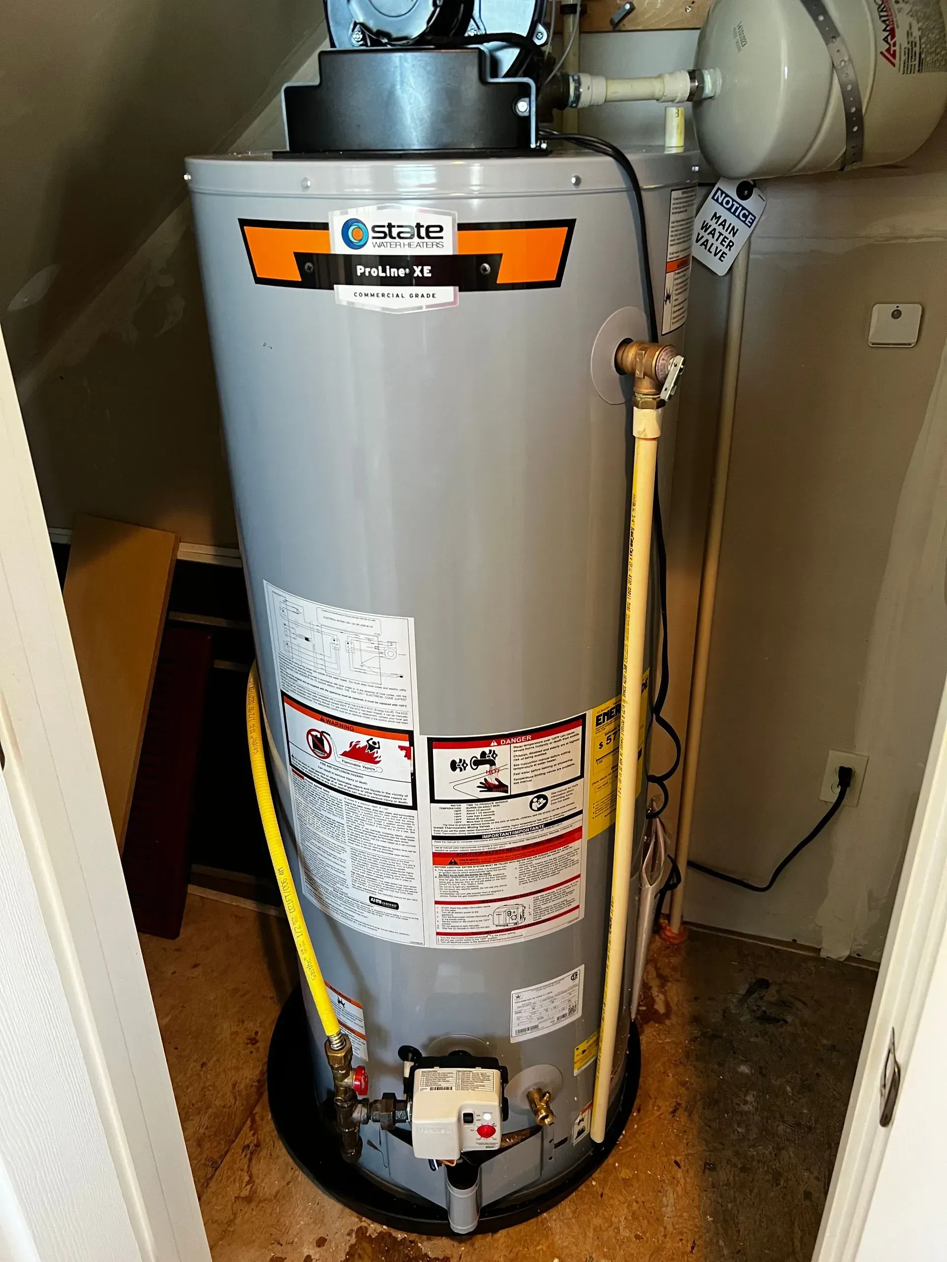 Gray water heater in a small utility closet with pipes, valves, and warning labels.