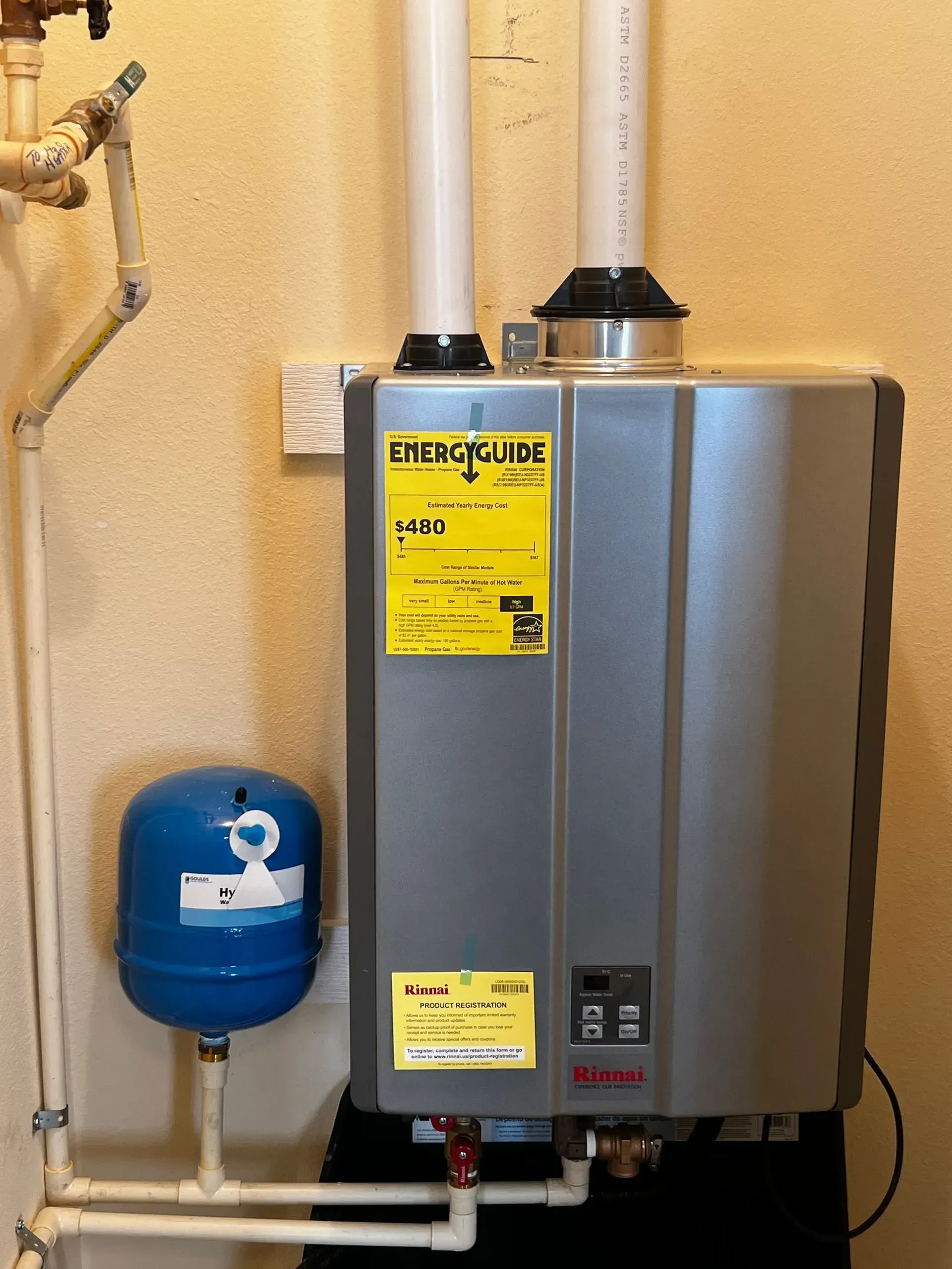 Wall-mounted gray tankless water heater with yellow labels beside a blue expansion tank in a utility room