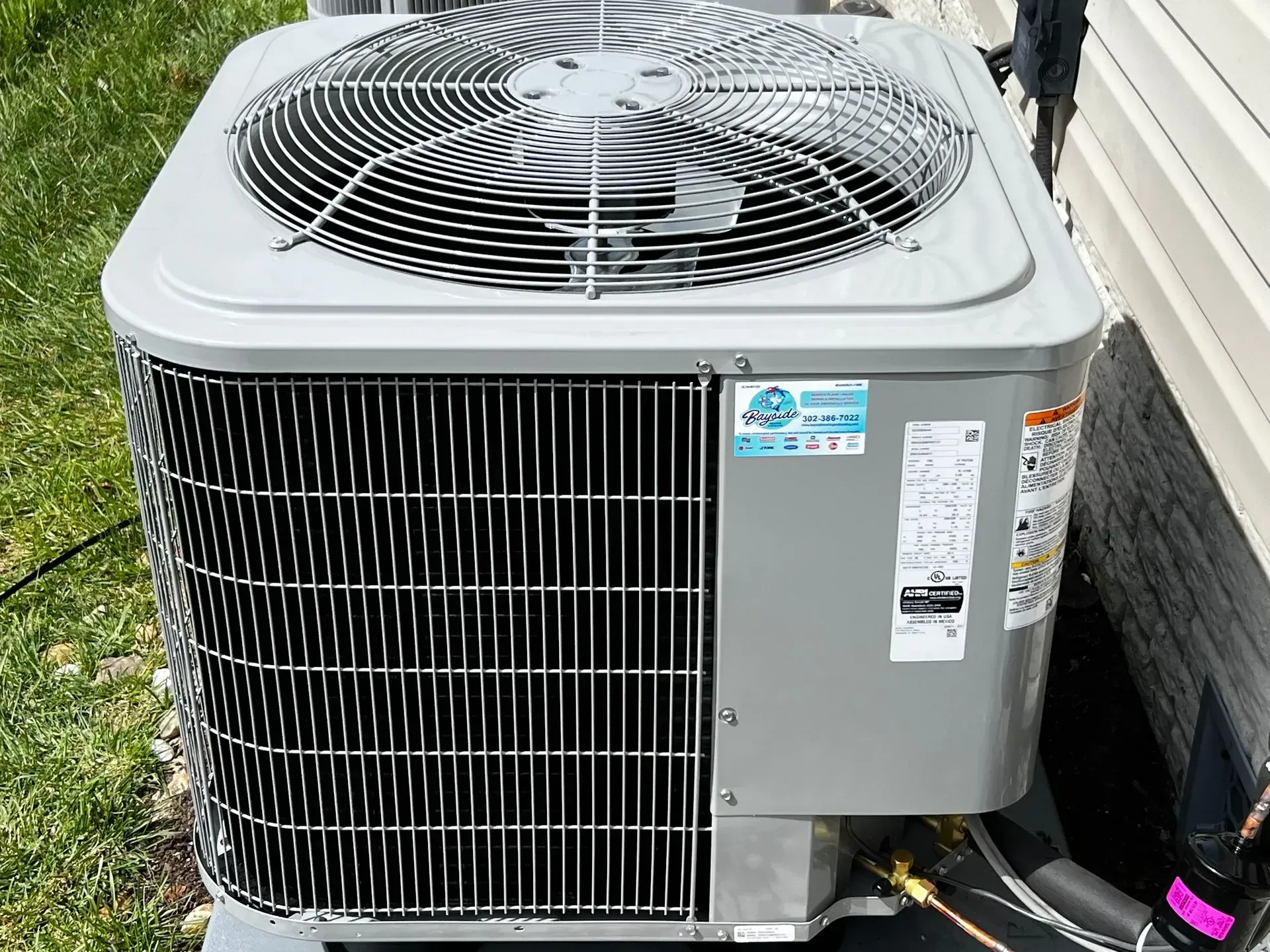 Outdoor air conditioner condenser unit beside a house, with fan grille and service labels.