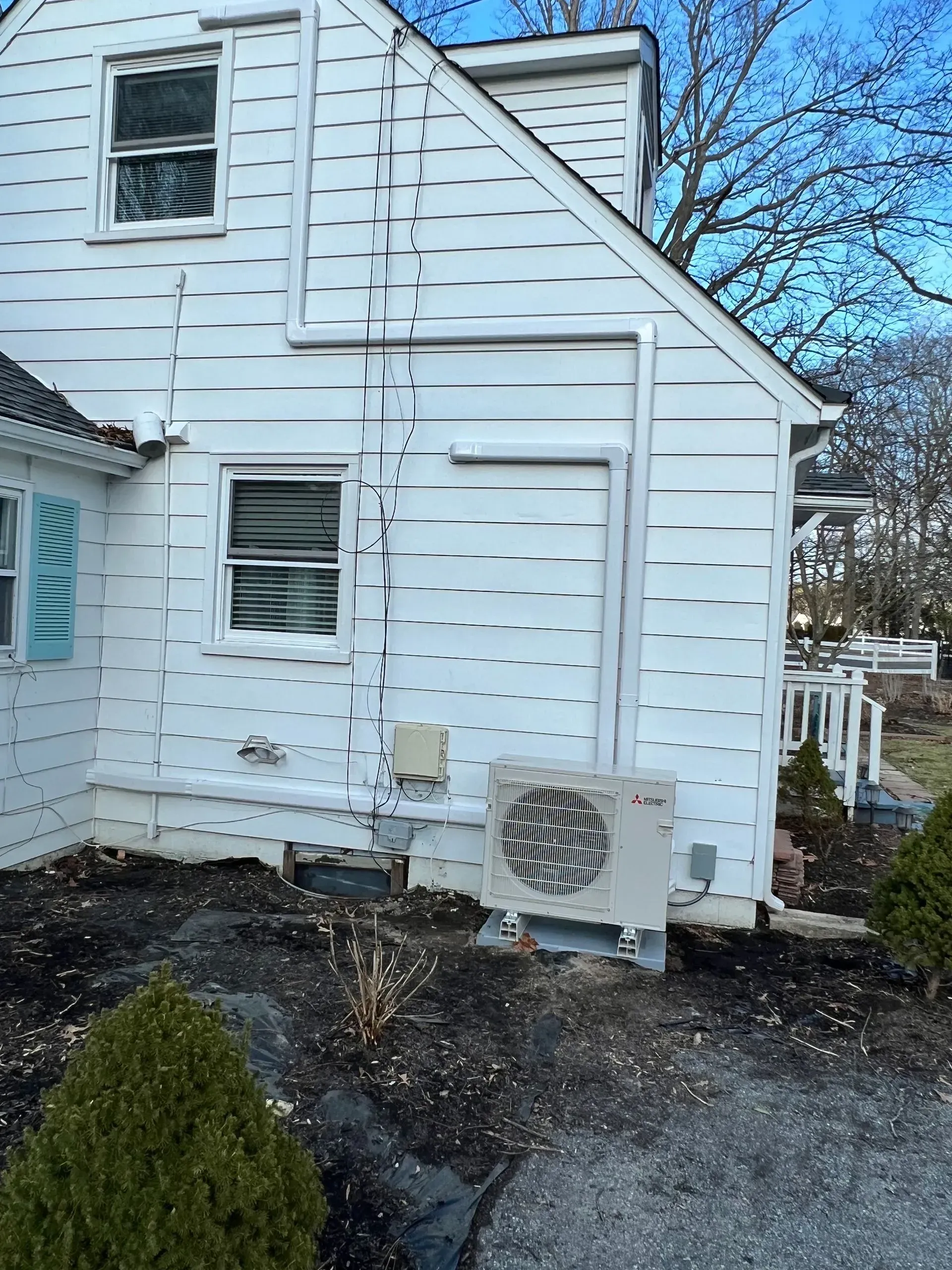 White house side with damaged siding and an outdoor AC unit beside a gravel driveway and shrubs