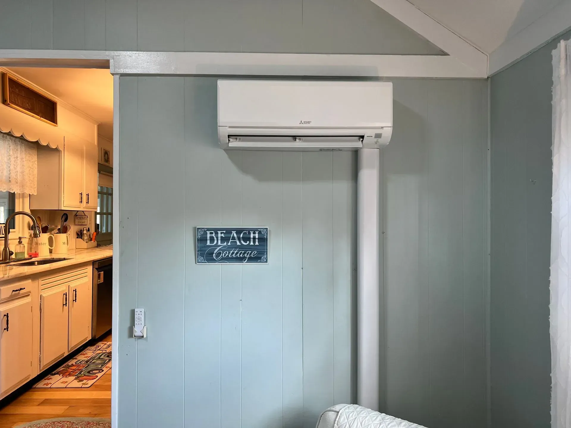 Small room with wall-mounted air conditioner and a blue “Beach” sign; kitchen visible through doorway.