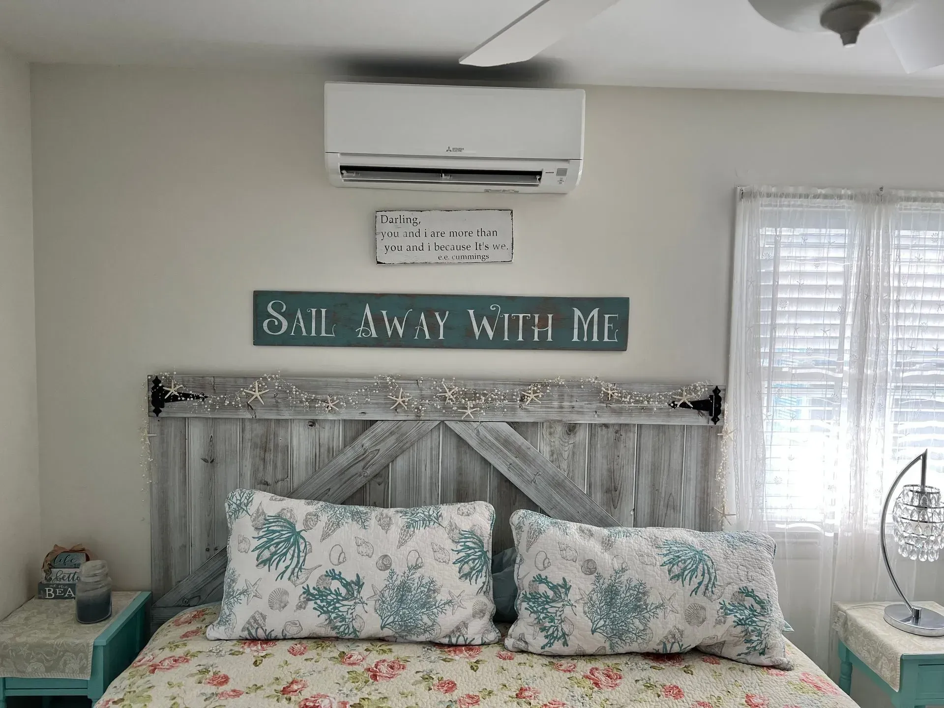 Cozy bedroom with rustic white bed, floral pillows, teal accents, and a wall sign reading “Sail Away With Me”