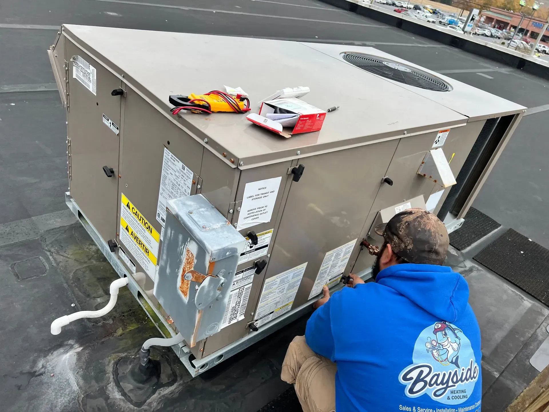 Technician kneeling beside rooftop HVAC unit, inspecting equipment with tools on wet pavement.