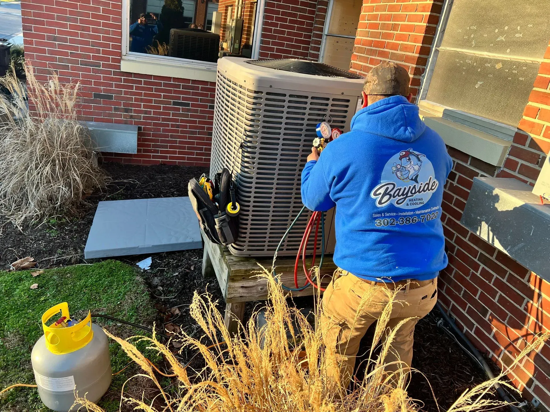 Worker in a blue hoodie services an outdoor HVAC unit beside a brick house.