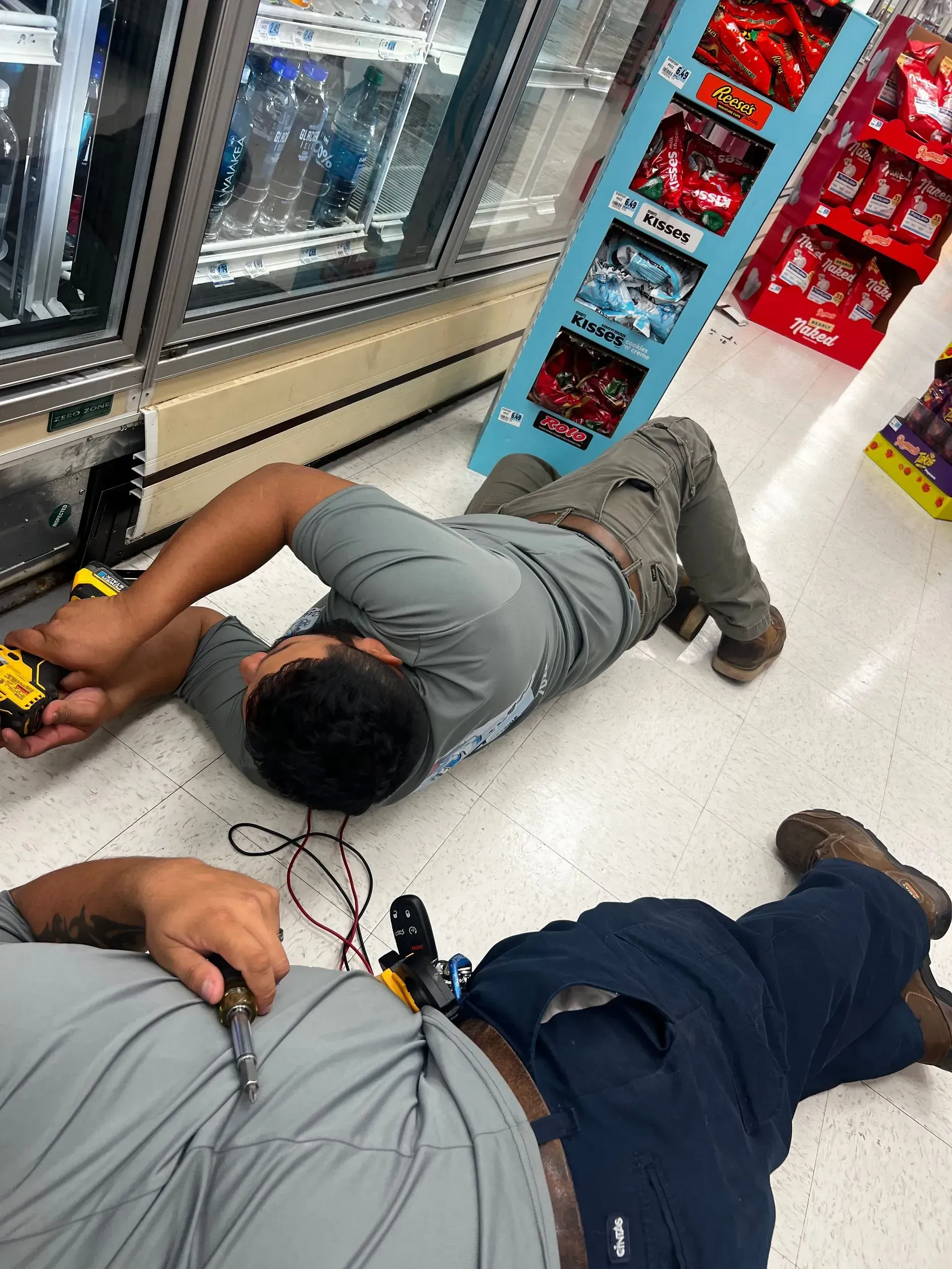 Two store employees lying on the floor near a refrigerated aisle, with cords and a red display nearby.